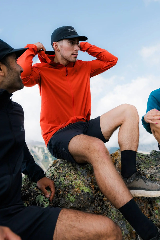 Man in red shirt and black shorts sitting on a rock with two other people, all outdoors.