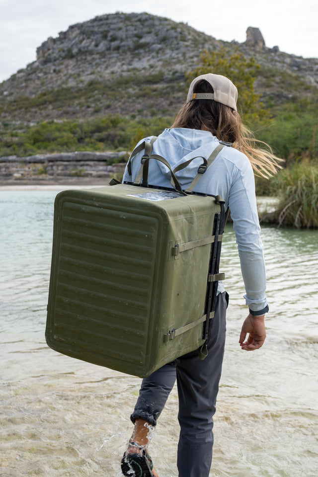 Person backpacking-carrying a RUX gear box by a body of water with mountains in the background
