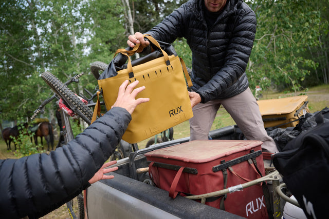 Two people handling a yellow RUX bag in an outdoor setting with trees and equipment.
