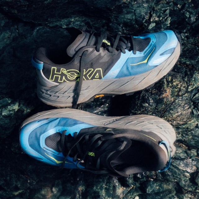 Pair of men's grey/blue Hoka Speedgoat 7 running shoes on rocky surface