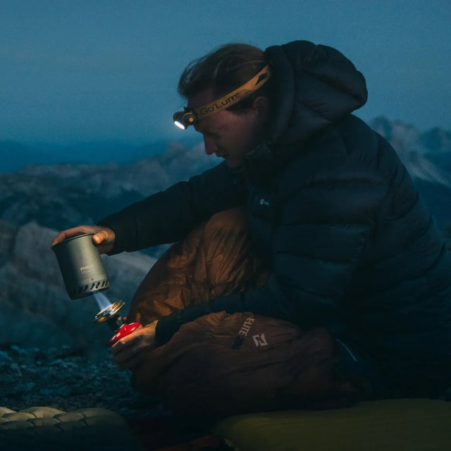 Person camping in the mountains at night, using a portable stove.