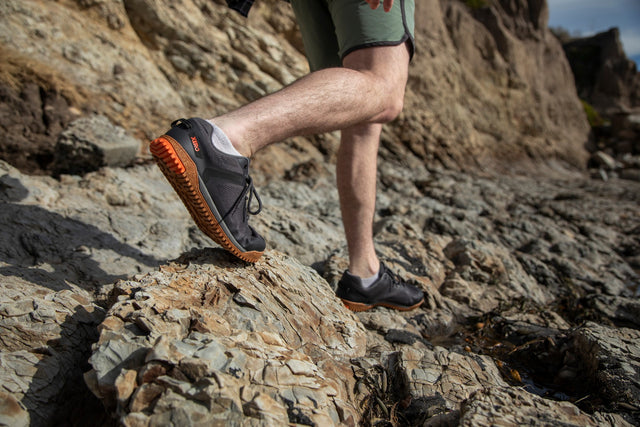 Person wearing black shoes with orange soles on a rocky surface