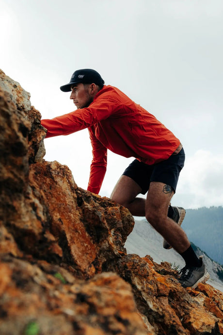 Person climbing a rocky mountain trail wearing a red jacket and black cap.