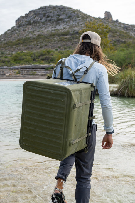 Person backpacking-carrying a RUX gear box by a body of water with mountains in the background