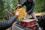 Two people handling a yellow RUX bag in an outdoor setting with trees and equipment.