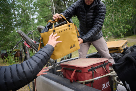 Two people handling a yellow RUX bag in an outdoor setting with trees and equipment.