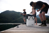Two people on a dock with fishing gear, one person opening a white RUX soft cooler bag