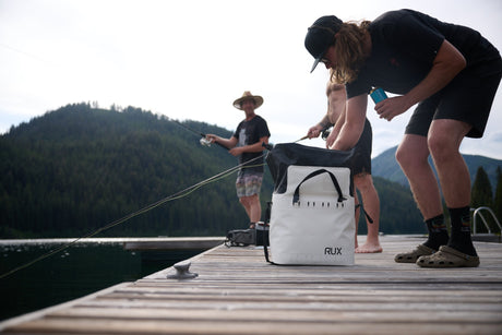 Two people on a dock with fishing gear, one person opening a white RUX soft cooler bag
