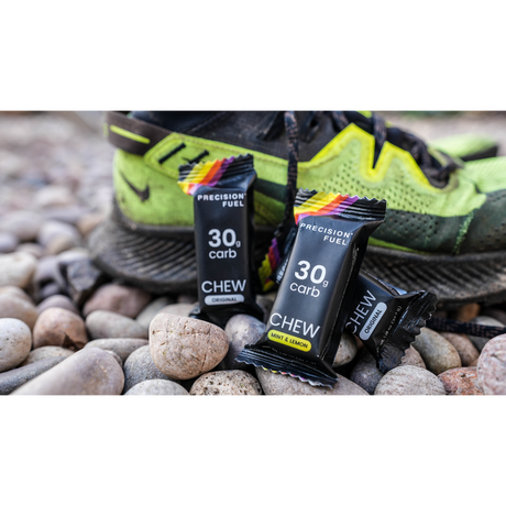 Two 'Precision Fuel' chew packets on pebbles with a running shoe in the background