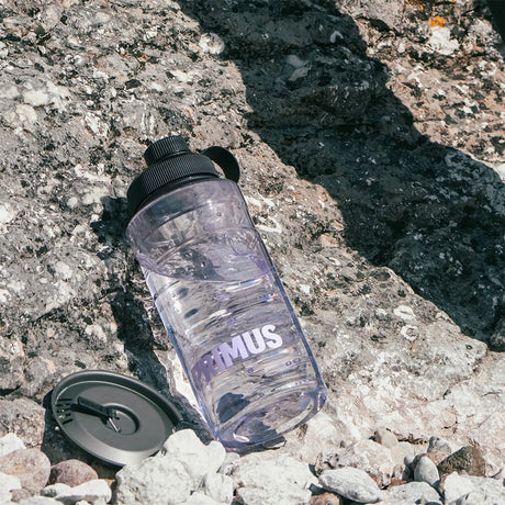 Clear water bottle with black cap on a rocky surface, featuring the Primus logo.
