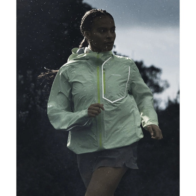 A woman running outdoors in the rain wearing a light green Salomon Bonatti Pro jacket