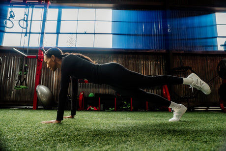 Person doing a plank exercise in a gym setting with large windows and gym equipment.