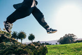 Person in black pants and shoes jumping over a log with palm trees and a clear sky in the background