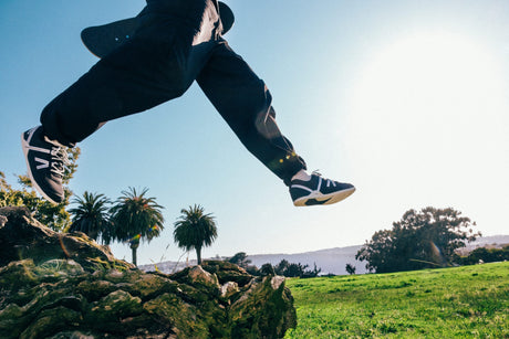 Person in black pants and shoes jumping over a log with palm trees and a clear sky in the background