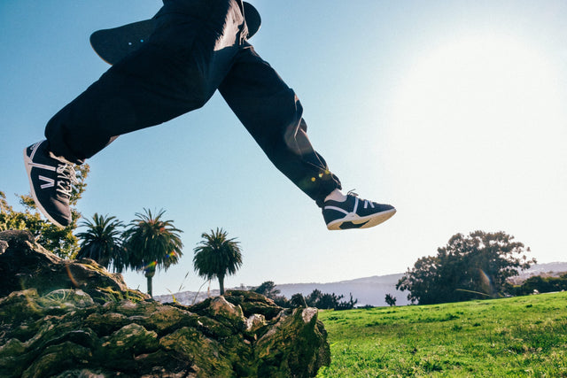 Person in black pants and shoes jumping over a log with palm trees and a clear sky in the background
